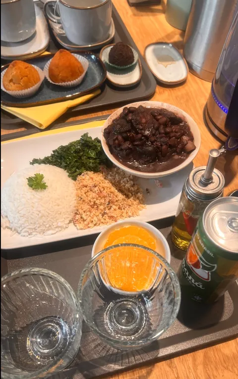 Feijoada-annos tarjottimella: papupataa kulhossa, valkoista riisiä, farofaa ja paahdettua lehtikaalia, sivussa appelsiinilohkoja ja guaraná-tölkki. Taustalla kaksi coxinhaa ja brigadeiro.