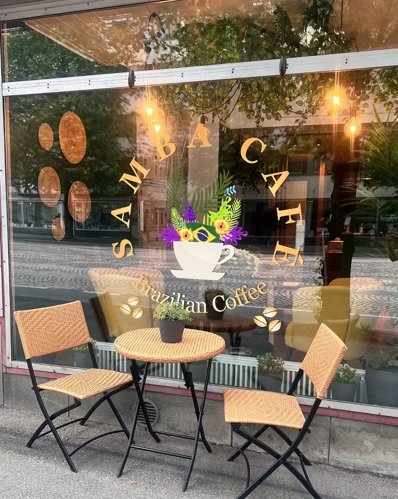 Samba Cafén katuikkuna, jossa kultainen Samba Café – Brazilian Coffee -logo. Ikkunan edessä rottinkinen bistropöytä ja kaksi tuolia.